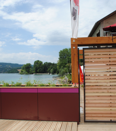 vue directe sur l'entre de la plage depuis la terrasse du snack. Petit muret et portail en bois ferm, vue du lac en arriere plan.