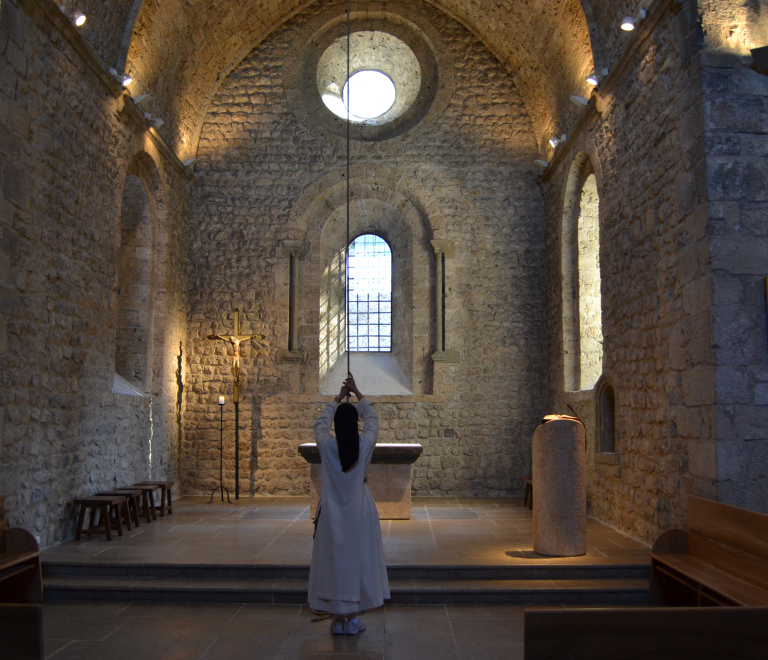 Au coeur de l'difice clair d'une lumire filtre par les vitraux, une personne en habit religieux s'apprte  tirer la ficelle relie  la cloche de l'glise